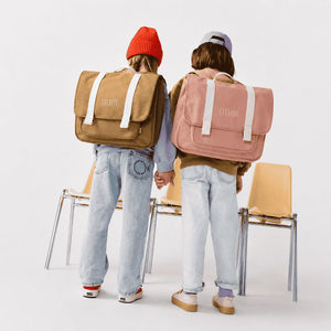 Two children seen from behind holding hands and wearing large canvas backpacks, one tan and one dusty pink, with the brand names 'GRATYI' and 'CAPRISI' printed on the back, standing in front of yellow chairs against a white studio background.