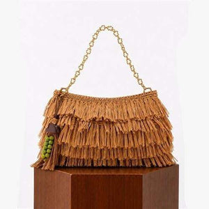 A tan woven fringe handbag with a gold chain handle and a green beaded tassel charm, displayed on a brown wooden pedestal against a white background.
