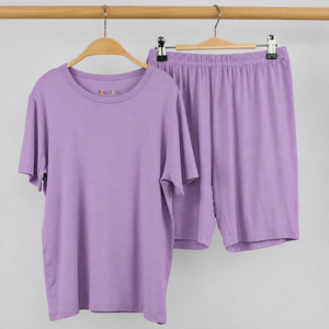 A lavender purple two-piece lounge set consisting of a short-sleeve crewneck t-shirt and matching shorts, hanging on wooden hangers from a metal clothing rack against a light gray wall.