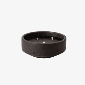 A dark brown textured ceramic candle holder containing a black wax candle with three white wicks, set against a plain white background.